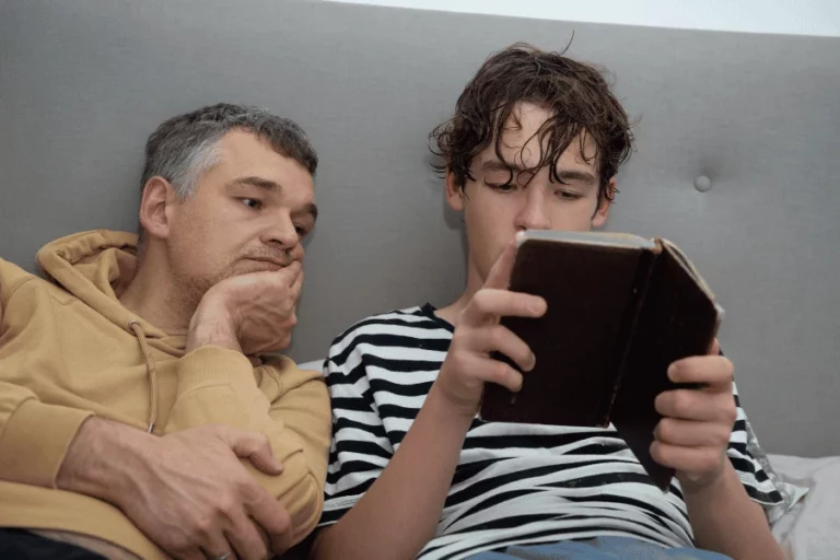 Vader zit half liggend op de bank en kijkt mee in het boek van zijn puberzoon. Een rustig moment van aandacht en betrokkenheid, waarin ruimte is voor contact en verbinding.