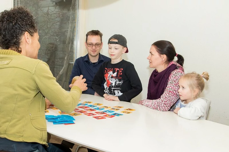 Gezin zit samen aan tafel met Fiona en gebruikt kwaliteitskaarten en een schudbol tijdens gezinsbegeleiding.