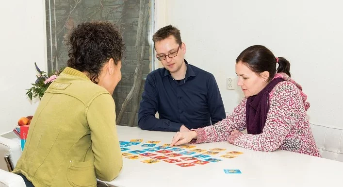 Kinder- en gezinscoach Fiona in gesprek met ouders aan tafel. Op tafel liggen de kwaliteitskaartjes die zij samen bespreken.