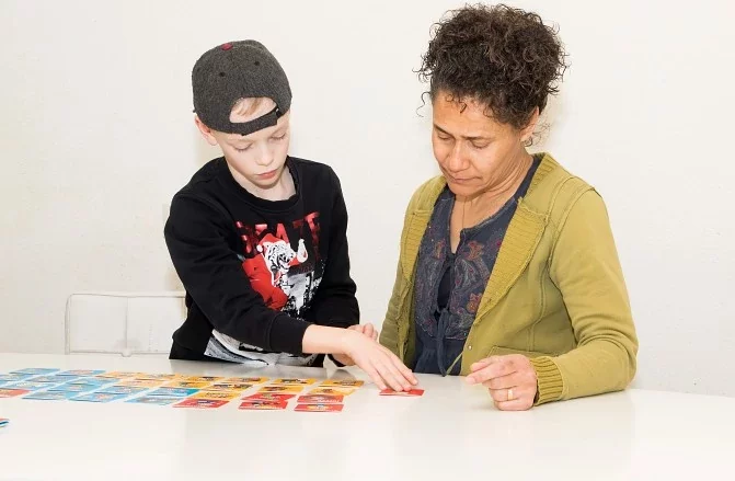 Een kinder- en gezinscoach zit naast een jongen aan tafel. Ze zijn samen in contact en spelen aan tafel, wat een ontspannen en betrokken sfeer uitstraalt.