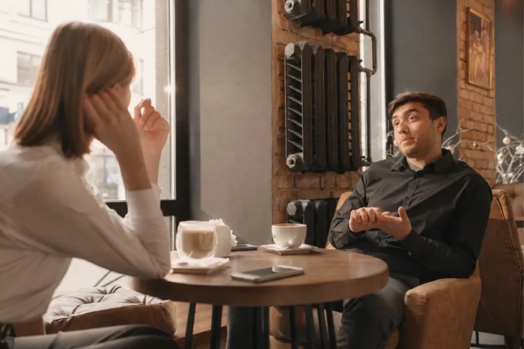 Een man en vrouw zitten ontspannen tegenover elkaar aan een rond tafeltje en zijn met elkaar in gesprek. De warme kleuren en open sfeer stralen rust en verbinding uit, passend bij het belang van respectvolle communicatie tussen ouders tijdens en na een scheiding.