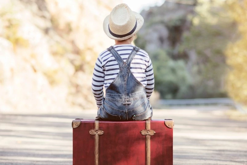 Een jongen zit met zijn rug naar de kijker op een koffer midden op een weg en kijkt vooruit. Het beeld symboliseert de onzekerheid en verwarring die een kind kan ervaren wanneer ouders uit elkaar gaan en het verlangt naar rust en vriendelijk contact tussen hen.