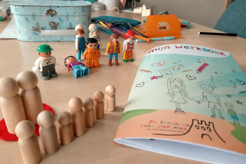 Het werkboek van het programma De Kinderen Scheiden Mee ligt op tafel, naast houten poppetjes en Playmobilfiguren die tijdens kindsessies worden gebruikt. De materialen helpen kinderen om gevoelens te herkennen, woorden te geven aan hun emoties en hun ervaringen rondom de scheiding op een veilige manier te uiten.